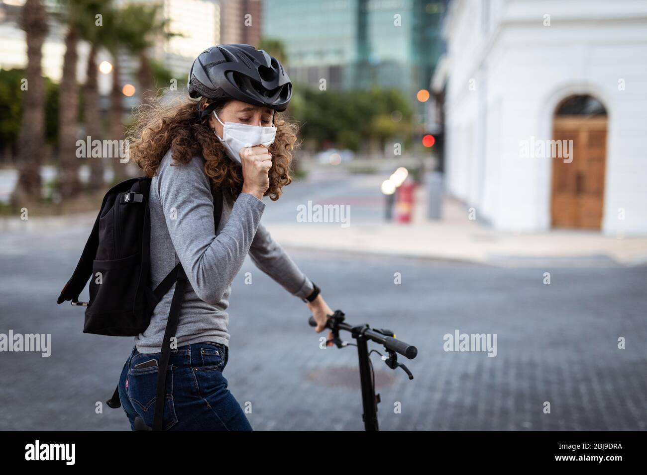 Kaukasische Frau, die auf der Straße eine Schutzmaske und einen Fahrradhelm trägt und auf der Straße hustet Stockfoto