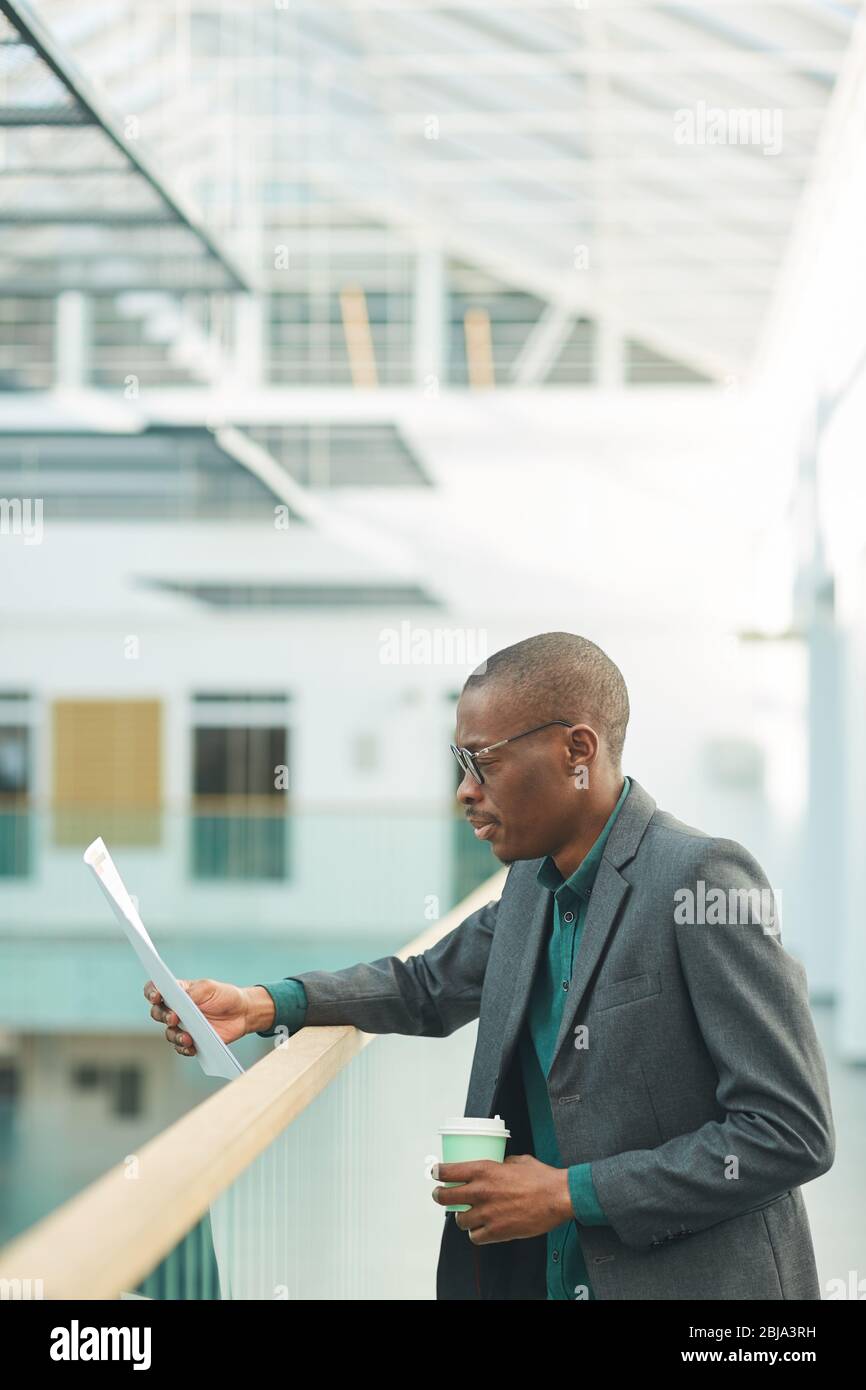 Afrikanischer junger Geschäftsmann, der Kaffee trinkt und ein Dokument liest, während er am Bürokorridor steht Stockfoto