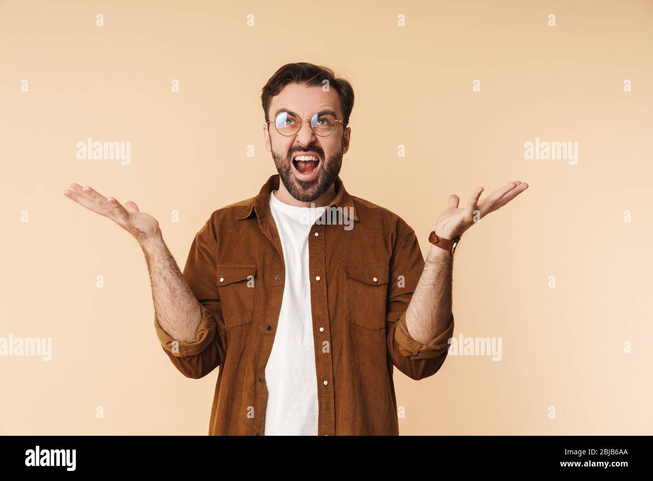 Porträt eines wütenden jungen, künstlerisch-bärtigen Mannes, der lässige Kleidung trägt, isoliert auf beigefarbenem Hintergrund stehend und schreiend Stockfoto