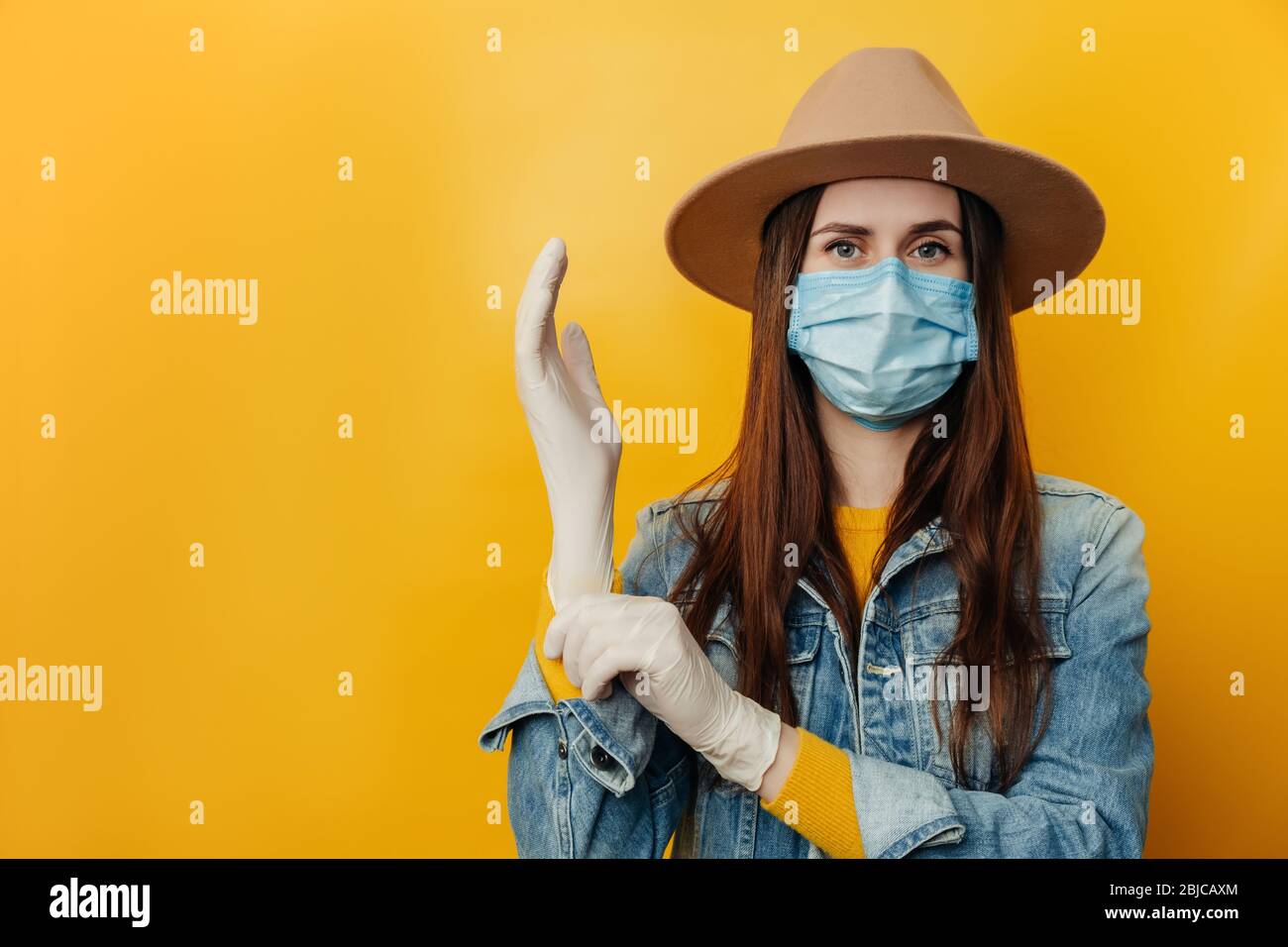 Attraktive junge Frau in Hut trägt schützende Gesichtsmaske, zieht medizinische Handschuhe an, Vorbereitung für eine gefährliche Reise während des Ausbruchs eines Coronavirus Stockfoto