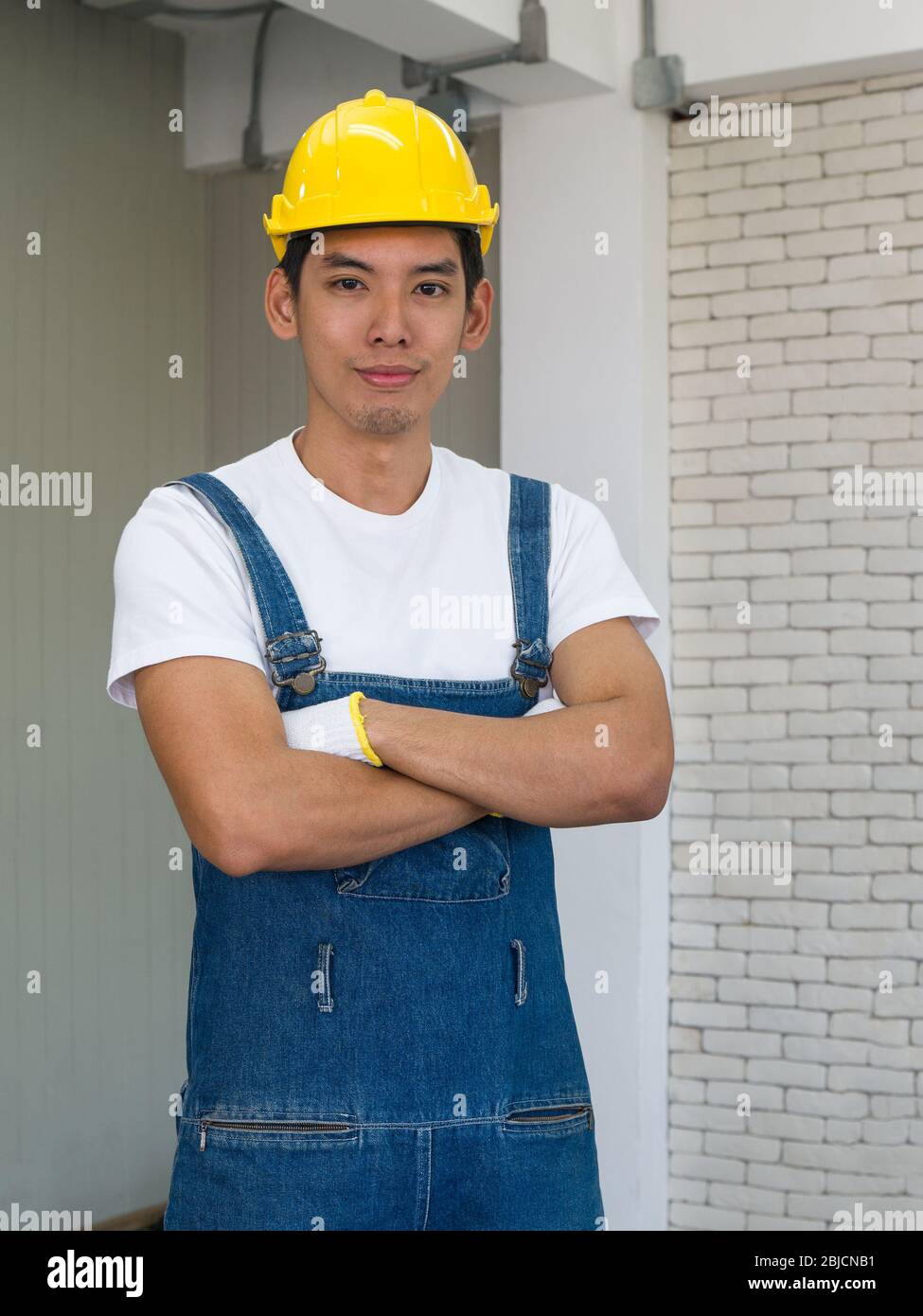 Ein asiatischer Zimmermann, der einen gelben Hut trägt, stellte sich im Werkstattzimmer mit den Armen. Stockfoto
