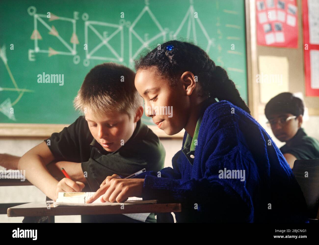 Junior Schule Schulmädchen schwarz African 9-11 Jahre im Klassenzimmer arbeiten an ihrem Schreibtisch mit blonde kaukasischen Jungen Schüler Geometrie Tafel hinter Stockfoto