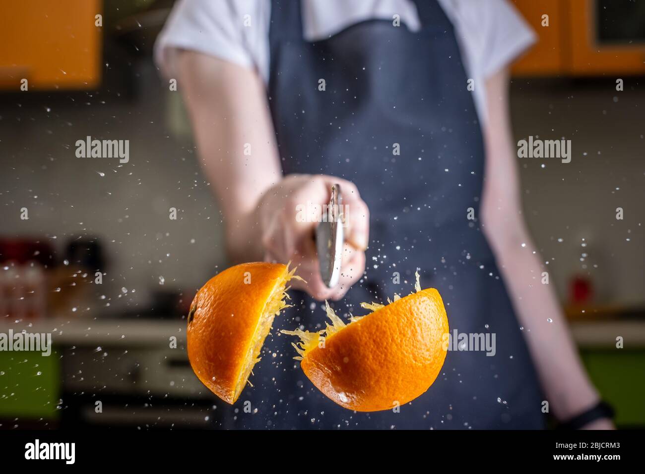 Koch schneidet eine Orange in zwei Hälften mit einem großen Messer in Bewegung. Wasser- und Saftspritzer fliegen in verschiedene Richtungen und frieren in der Luft ein Stockfoto