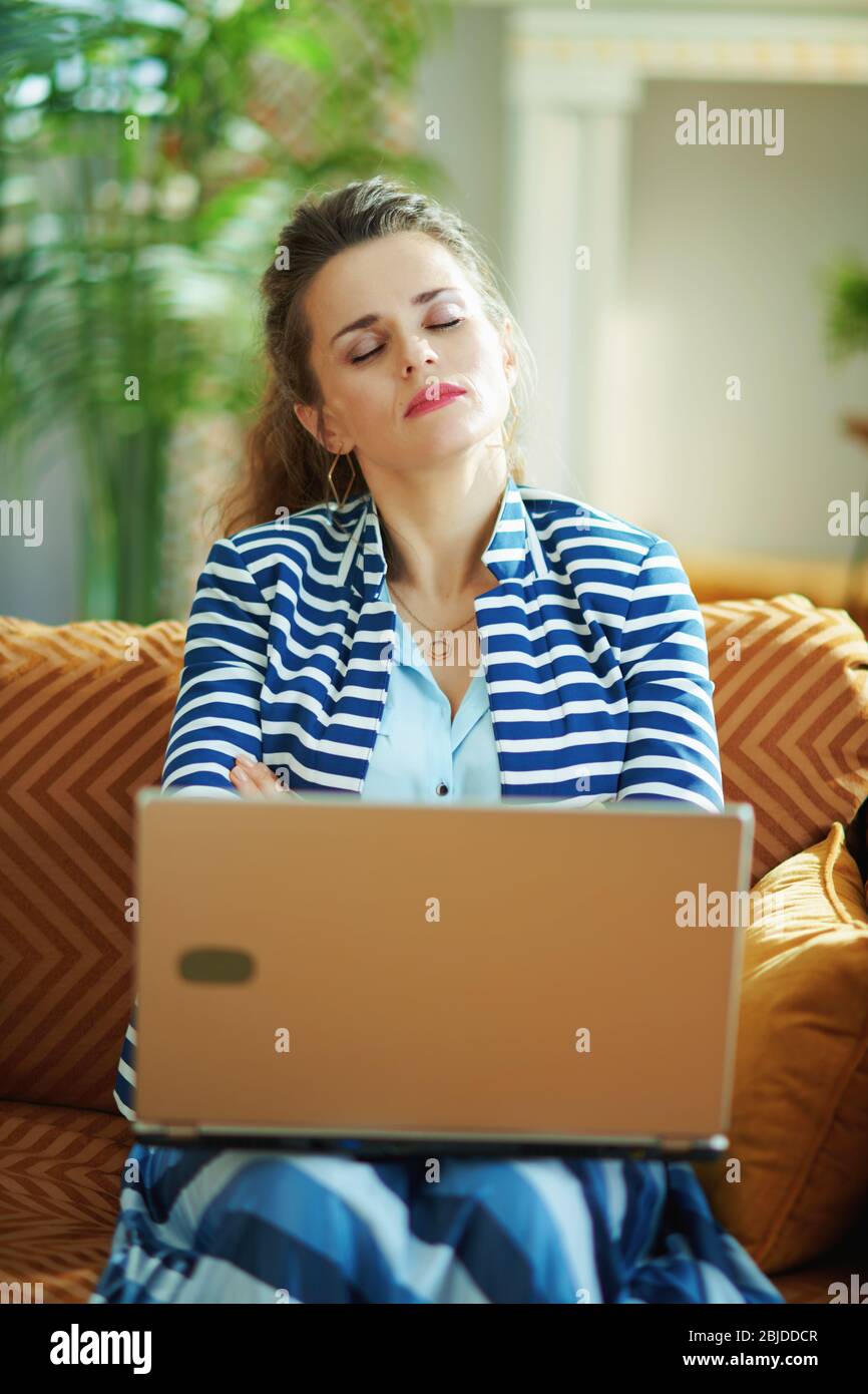 Gestresste moderne 40 Jahre alte Hausfrau in blauer Bluse und gestreiften Jacke sitzt auf Diwan mit Laptop im modernen Haus an sonnigen Tag. Stockfoto