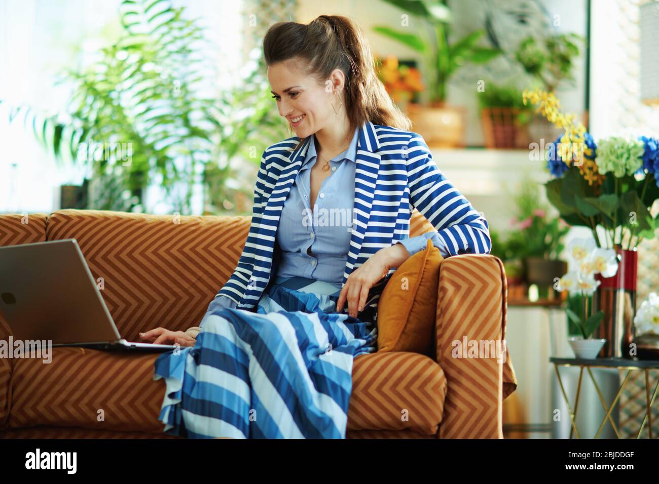 Glückliche junge Hausfrau in blauer Bluse und gestreiften Jacke sitzt auf Diwan mit Laptop im modernen Wohnzimmer bei schönem Tag. Stockfoto