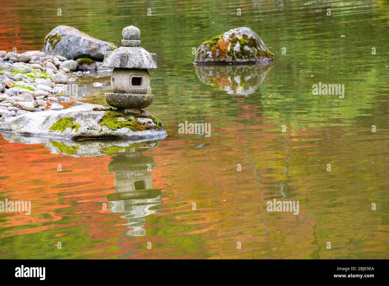 Japanischer Garten Steinlaterne & Rock im Teich mit verträumten Orange & Grün Reflexionen in Wellen, die den Teich bedecken Stockfoto