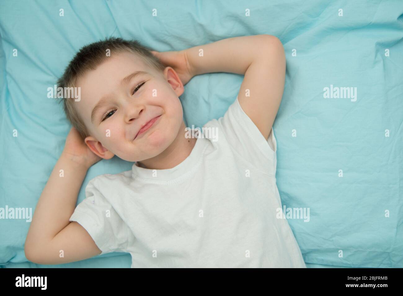 Der kleine Junge wacht in seinem Bett auf und lächelt. Kleinkind im weißen Hemd schaut zur Kamera Stockfoto