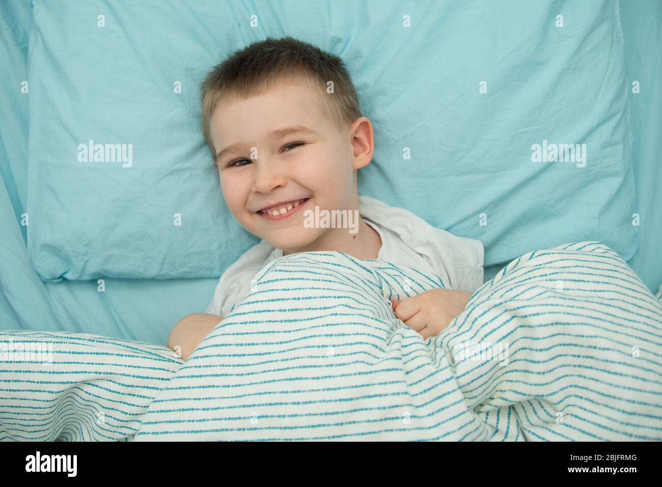 Der kleine Junge liegt in seinem Bett und lächelt. Kleinkind im weißen Hemd Stockfoto