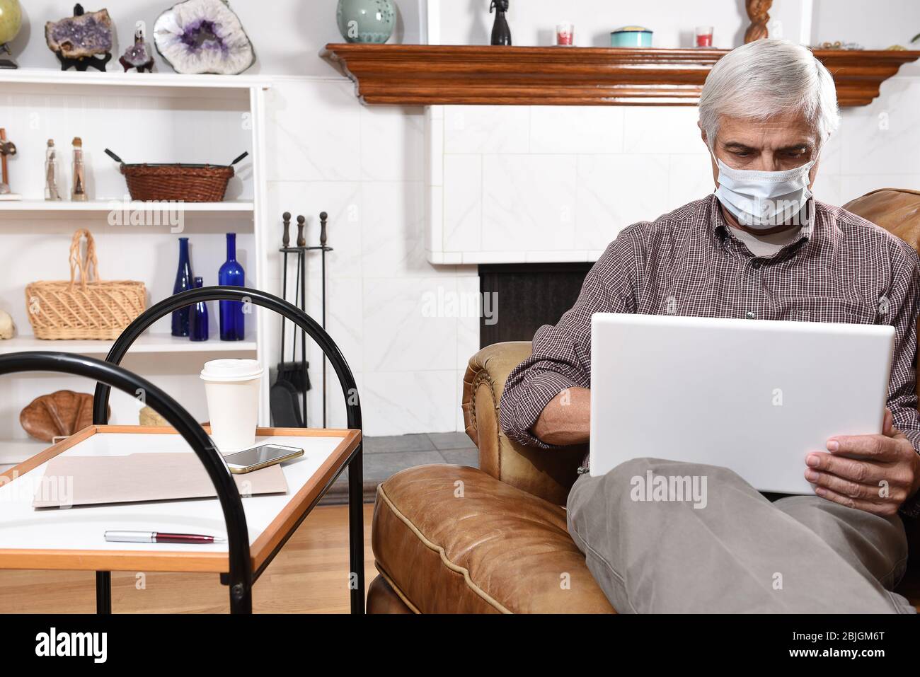 Ein reifer Mann, der während der COVID-19-Beschränkungen für den Aufenthalt zu Hause eine chirurgische Maske trägt und von zu Hause aus mit seinem Laptop und Handy arbeitet. Stockfoto