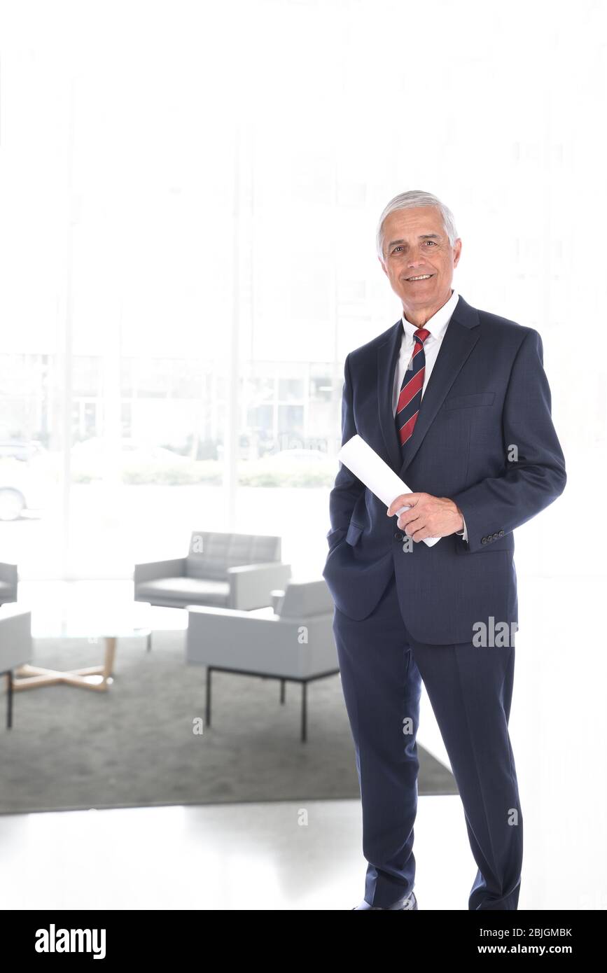 Lächelnder, munter Geschäftsmann in einer modernen Lobby. Der Mensch hält ein zusammengerolltes Dokument in der Hand. Vertikales Format mit High Key Backg Stockfoto