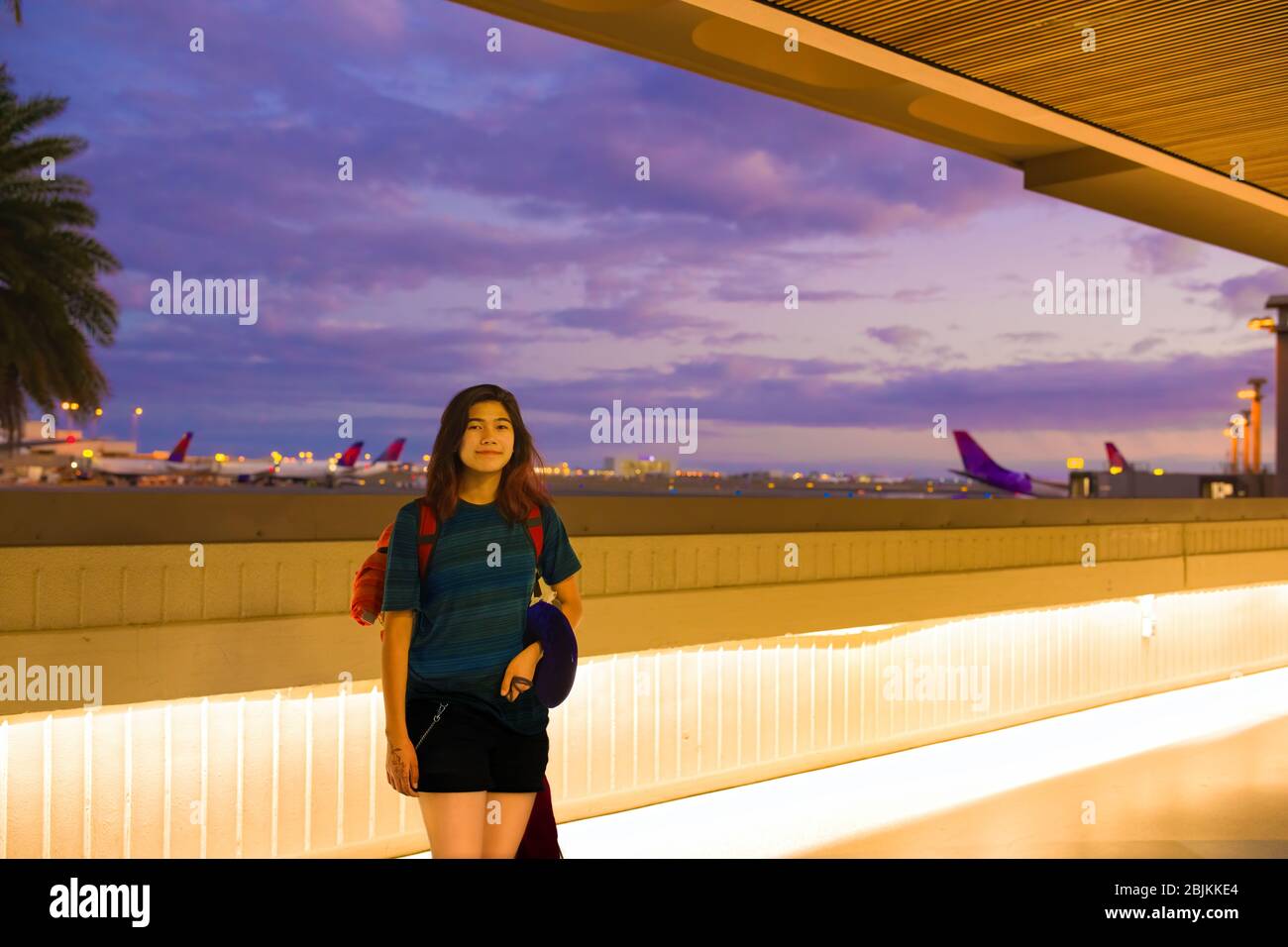 Teenager-Mädchen mit rotem Rucksack, das am Außenkorridor des hawaiianischen Flughafens steht, bevor sie am Abend abfliegt. Flugzeuge und bunte Himmel im Bac Stockfoto