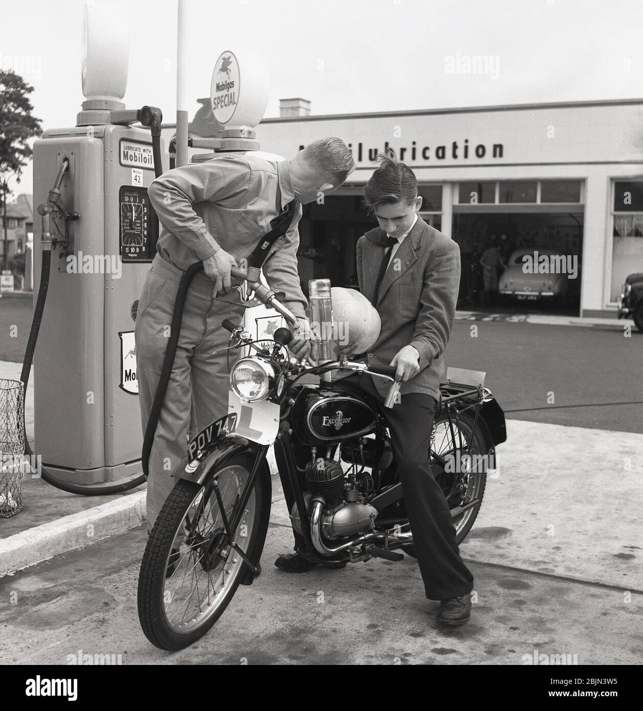 1950s, historisch, ein junger Mann, der auf einem britischen Excelsior Motorrad draußen in einer Garage mit Benzin in seinem Tank von einem uniformierten Vorplatz Aufseher, England, Großbritannien. Excelsior mit Sitz in Coventry war Großbritanniens erster Motorradhersteller, der 1896 sein eigenes 'Motorfahrrad' hergestellt hatte. Stockfoto
