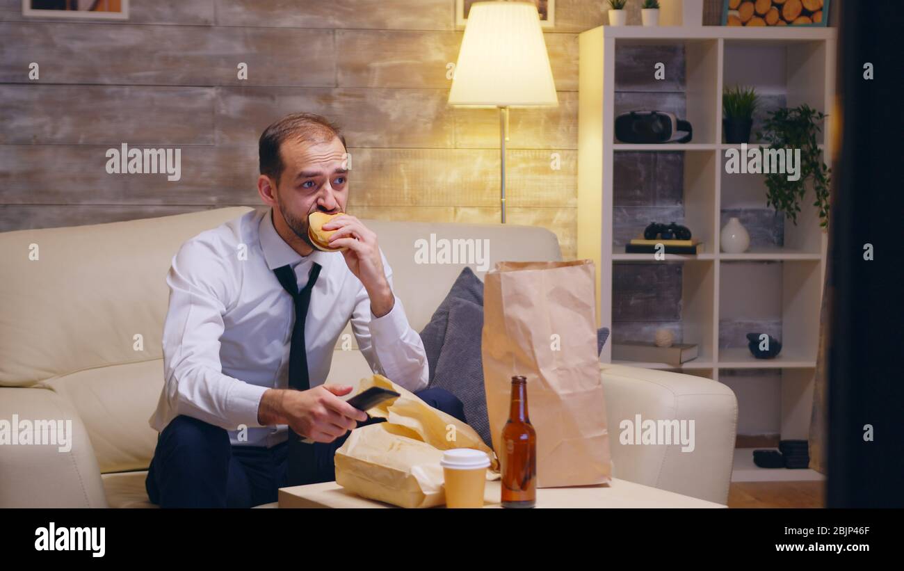 Kaukasischer Geschäftsmann lösen seine Krawatte auf der Couch sitzen nach einem langen Tag bei der Arbeit. Eating Burger. Stockfoto