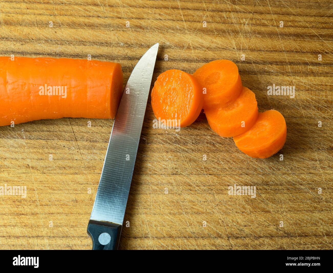 Frische Karotten und vier Karottenscheiben auf einem Holzhackbrett mit einem scharfen Küchenmesser Stockfoto