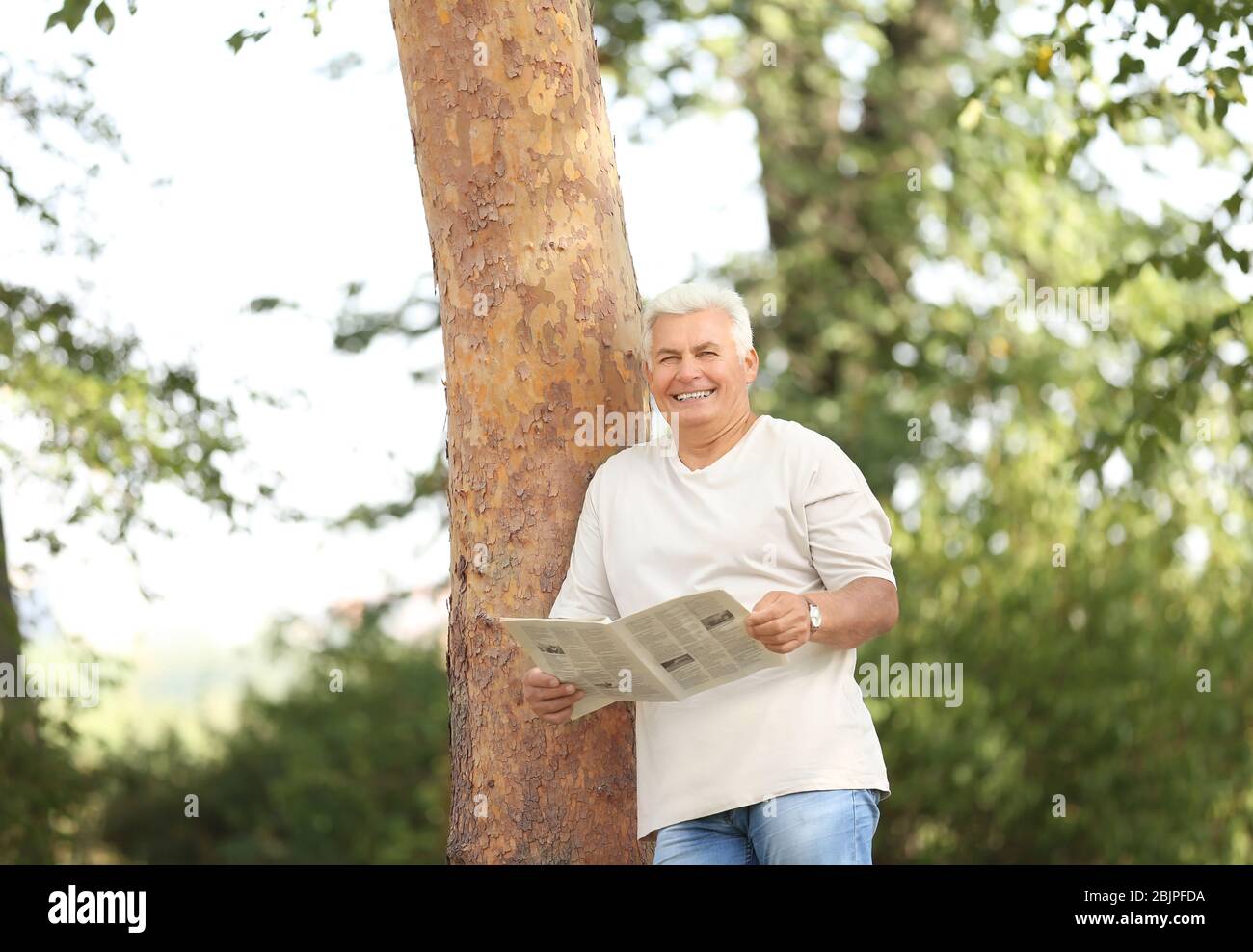 Schöner reifer Mann liest Zeitung in der Nähe Baum im Park Stockfoto