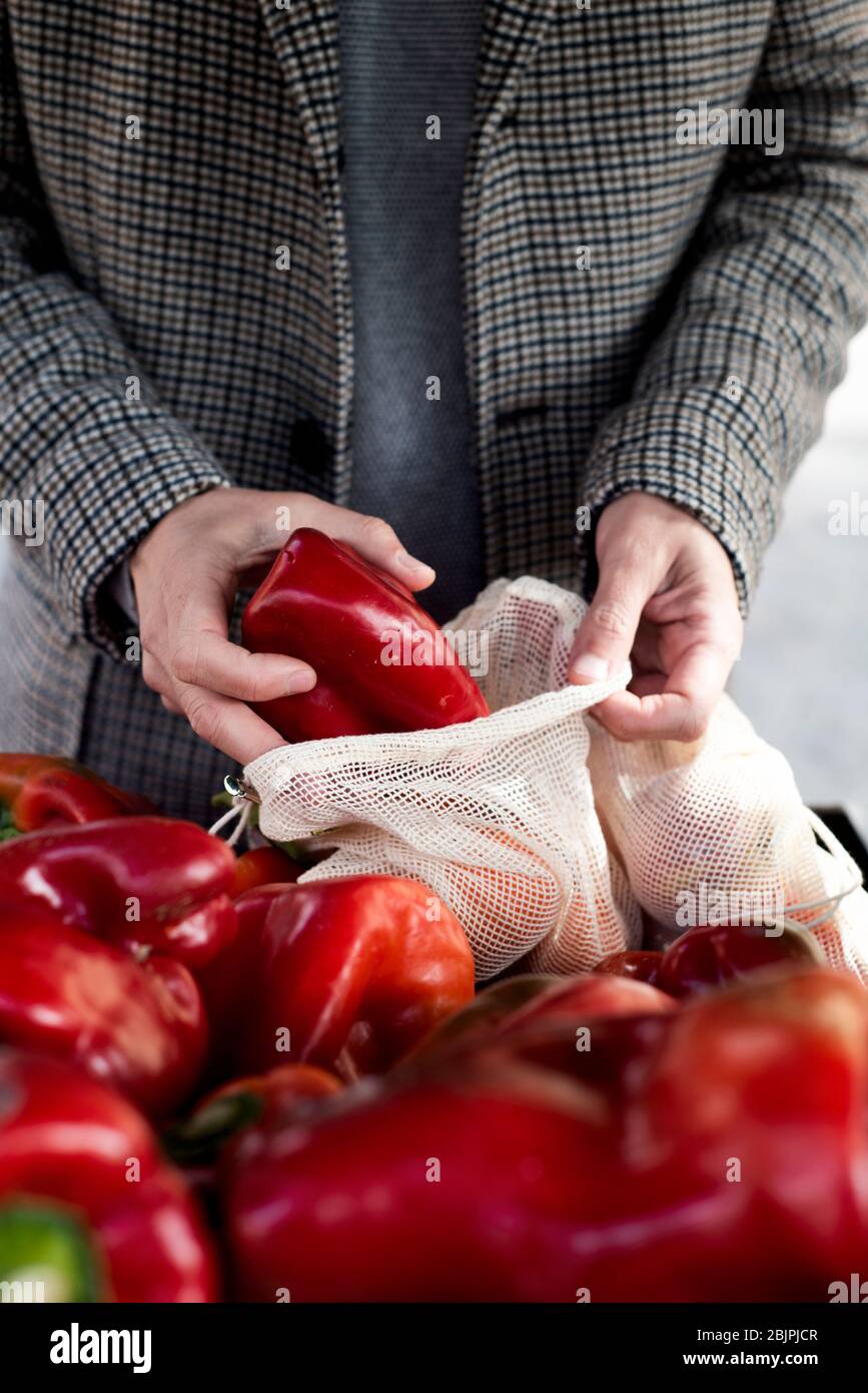 Nahaufnahme eines Mannes, der in einem Greengrocker einkauft und einige rote Paprika in einen wiederverwendbaren Textil-Netzbeutel stellt, um die Kunststoffverschmutzung zu verringern Stockfoto