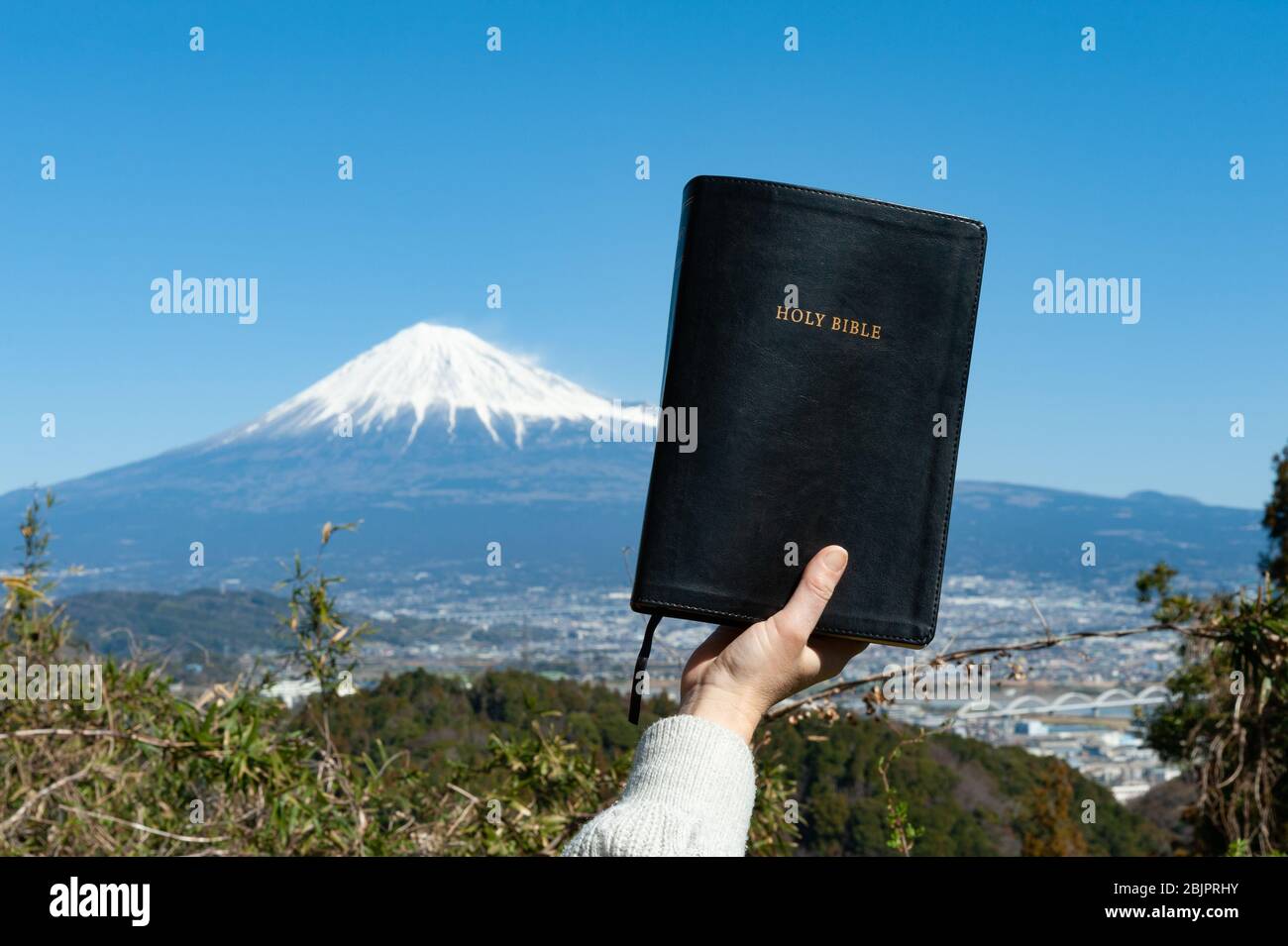 Erhobene Hand, die die Heilige Bibel hält. Hintergrund mit Luftaufnahme von Fuji City und Mount Fuji mit blauem Himmel an einem schönen Wintermorgen. Stockfoto