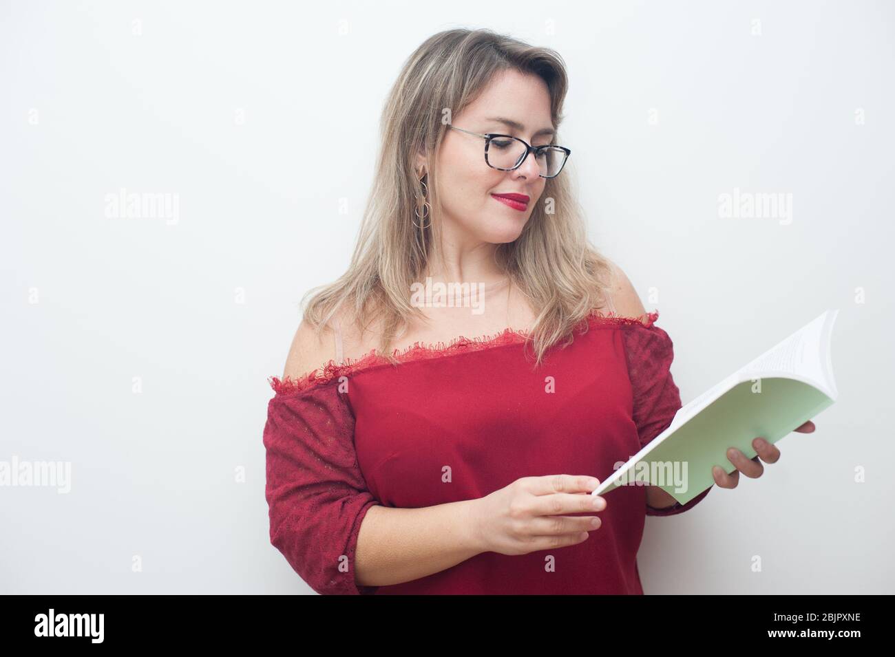 Lächelnde junge blonde Frau, die ein Buch in einem weißen Hintergrund liest. Sie trägt ein rotes Hemd und eine Brille. Großer Kopierplatz. Stockfoto