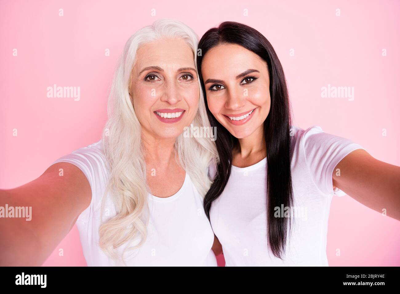 Nahaufnahme Foto von fröhlichen gealterten Mutter junge Tochter Damen machen Selfies besten Freunde gemütliche Atmosphäre tragen weiße T-Shirts isoliert pastellrosa Farbe Stockfoto