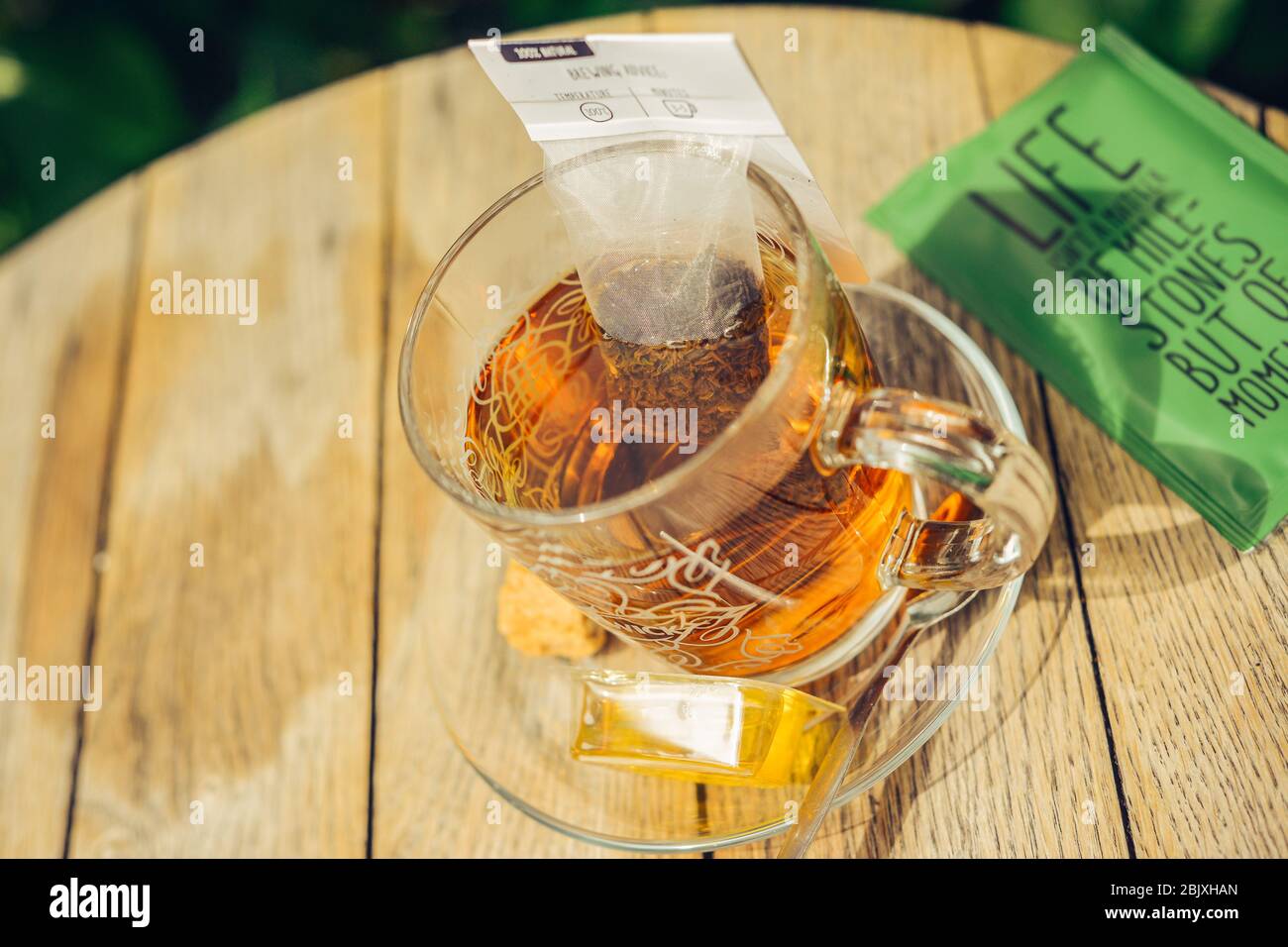 Nahaufnahme von Tee in Glas Tasse mit Teebeutel, Honig, Plätzchen, Zucker. Rustikaler Holztisch mit runder Tischuntergrund. Sonnenlicht im Freien. Lifestyle. Lebenszitat. Stockfoto