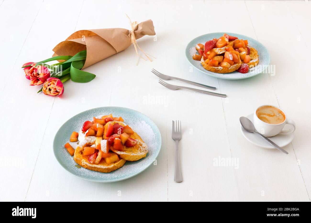 Französischer Toast mit geschmorten Äpfeln und Erdbeeren, mit einer Tasse Kaffee und Tulpenblumen auf einem weißen Holztisch. French Toast oder auch Pain perdu o Stockfoto