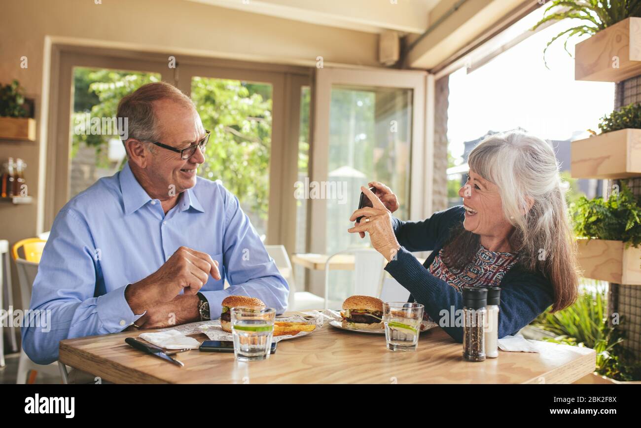 Lächelnde ältere Frau, die in einem Restaurant Fotos von ihrem Mann machte. Rentnerpaar genießen in einem Café Picutres mit einem Handy klicken. Stockfoto