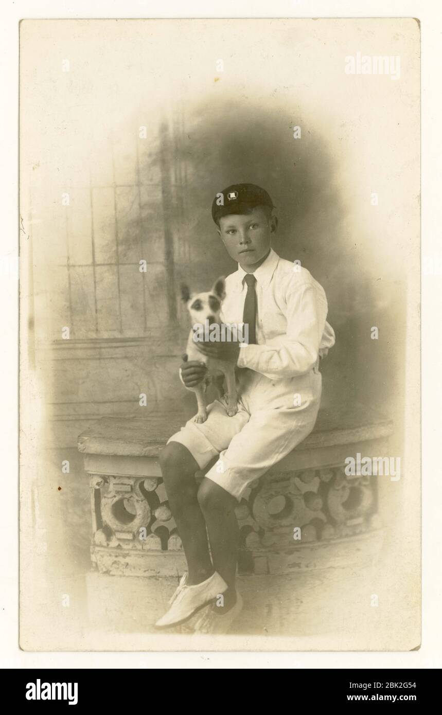 Postkarte des Jungen aus dem frühen 1900, der Mütze, Shorts, Cricket-Weiße trägt, einen Terrier in der Hand, London, Großbritannien um 1918 Stockfoto