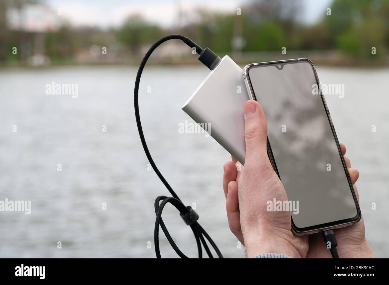Man benutzt Smartphone mit leerem weißen Bildschirm, während man von der Powerbank am Ufer des Flusses lädt. Modernes Technologiekonzept. Selektiver Fokus. Stockfoto