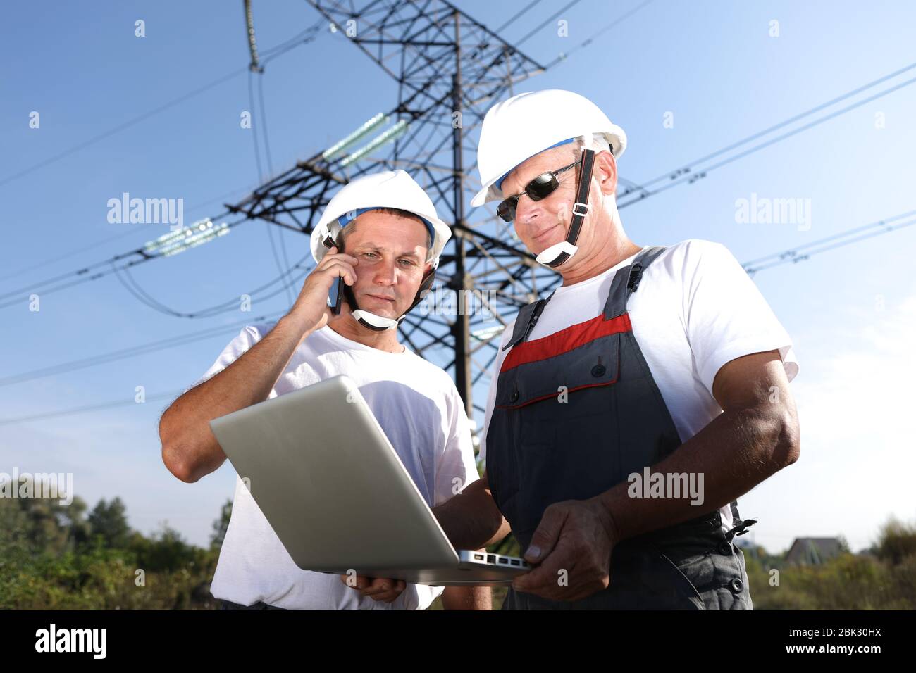 Zwei männliche Ingenieure mit einem Laptop auf einem Hintergrund von elektrischen Pylonen. Notfalldienst für die Reparatur von elektrischen Netzwerken. Ein Mann spricht am Telefon Stockfoto