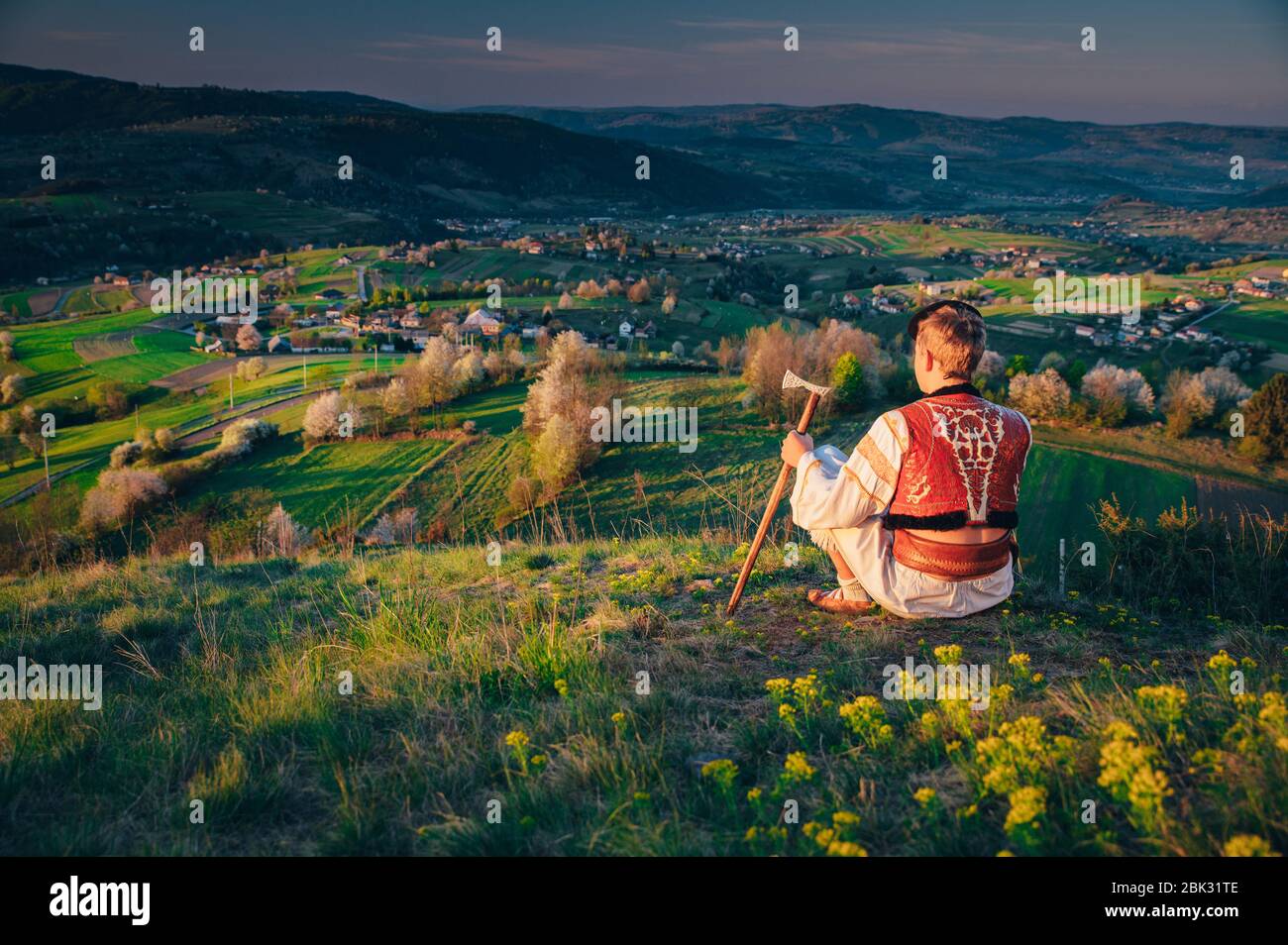 Mann in traditioneller Tracht sitzt in der Frühlingsnatur bei Sonnenaufgang. Bearbeiten Sie den Bereich. Schöne Frühlingslandschaft im Hintergrund. Hrinova Dorf in Slowakisch Stockfoto