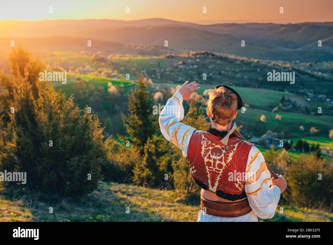 Mann in traditioneller Volkskleid, der das Licht des Sonnenaufgangs in einer wunderschönen, warmen Frühlingslandschaft betrachtet. Ländliche Wiese und Frühlingsblüten Kirschbäume im Hintergrund Stockfoto