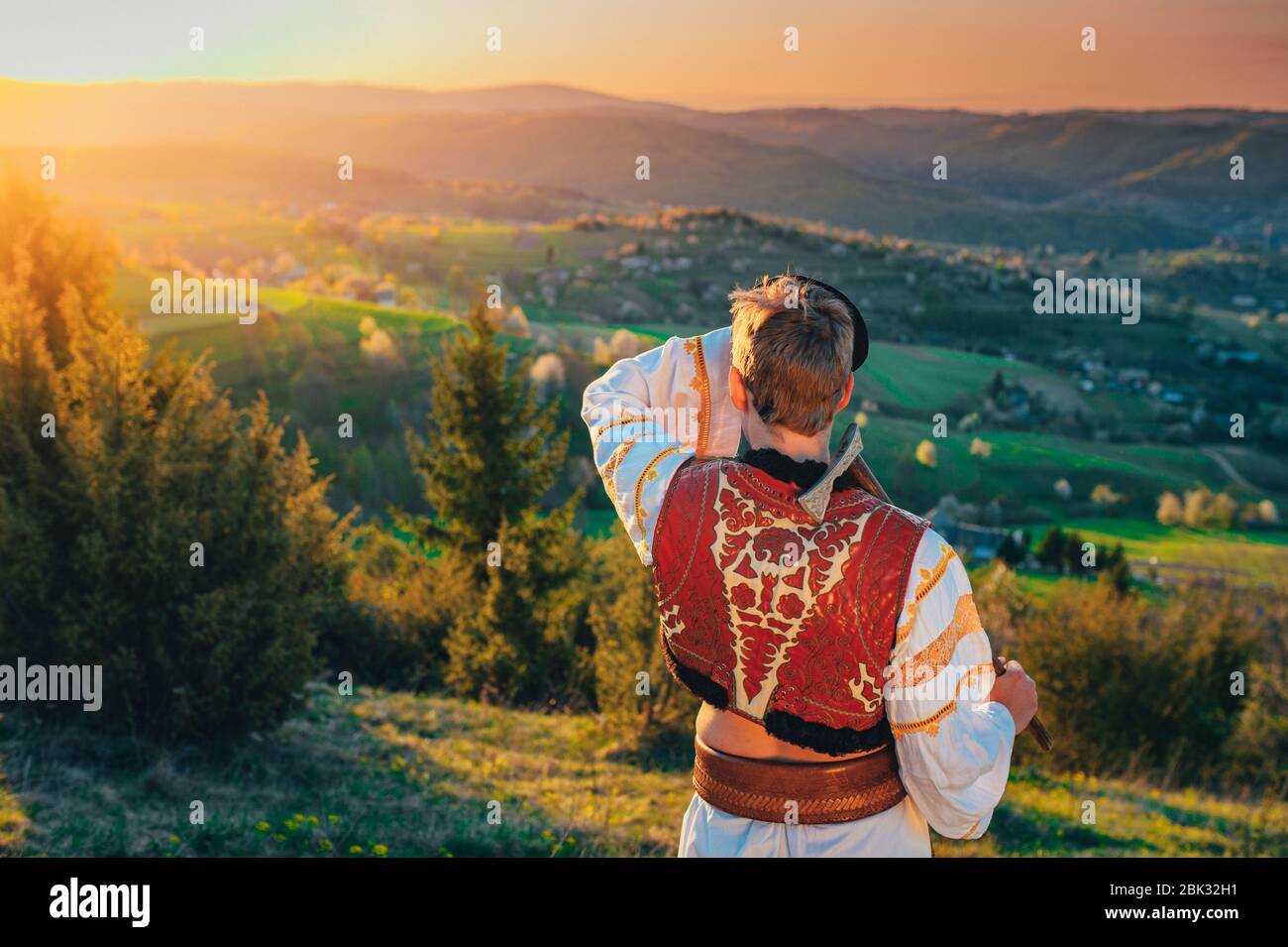 Ein junger Mann in slowakischer Tracht blickt auf die Frühlingslandschaft im Dorf Hrinova in der Slowakei. Aufgehende Sonne und Frühling blühende Bäume in der Stockfoto