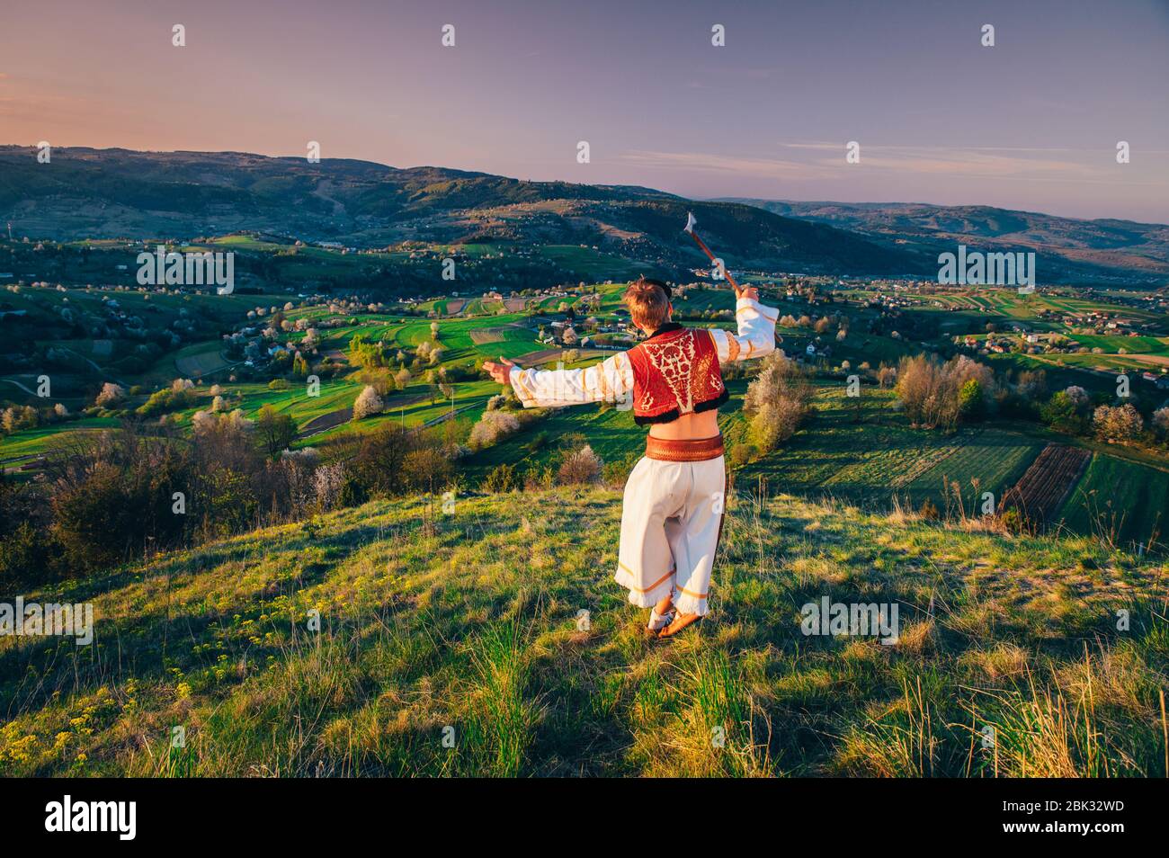 Ein junger Mann in slowakischer Tracht blickt auf die Frühlingslandschaft im Dorf Hrinova in der Slowakei. Aufgehende Sonne und Frühling blühende Bäume in der Stockfoto