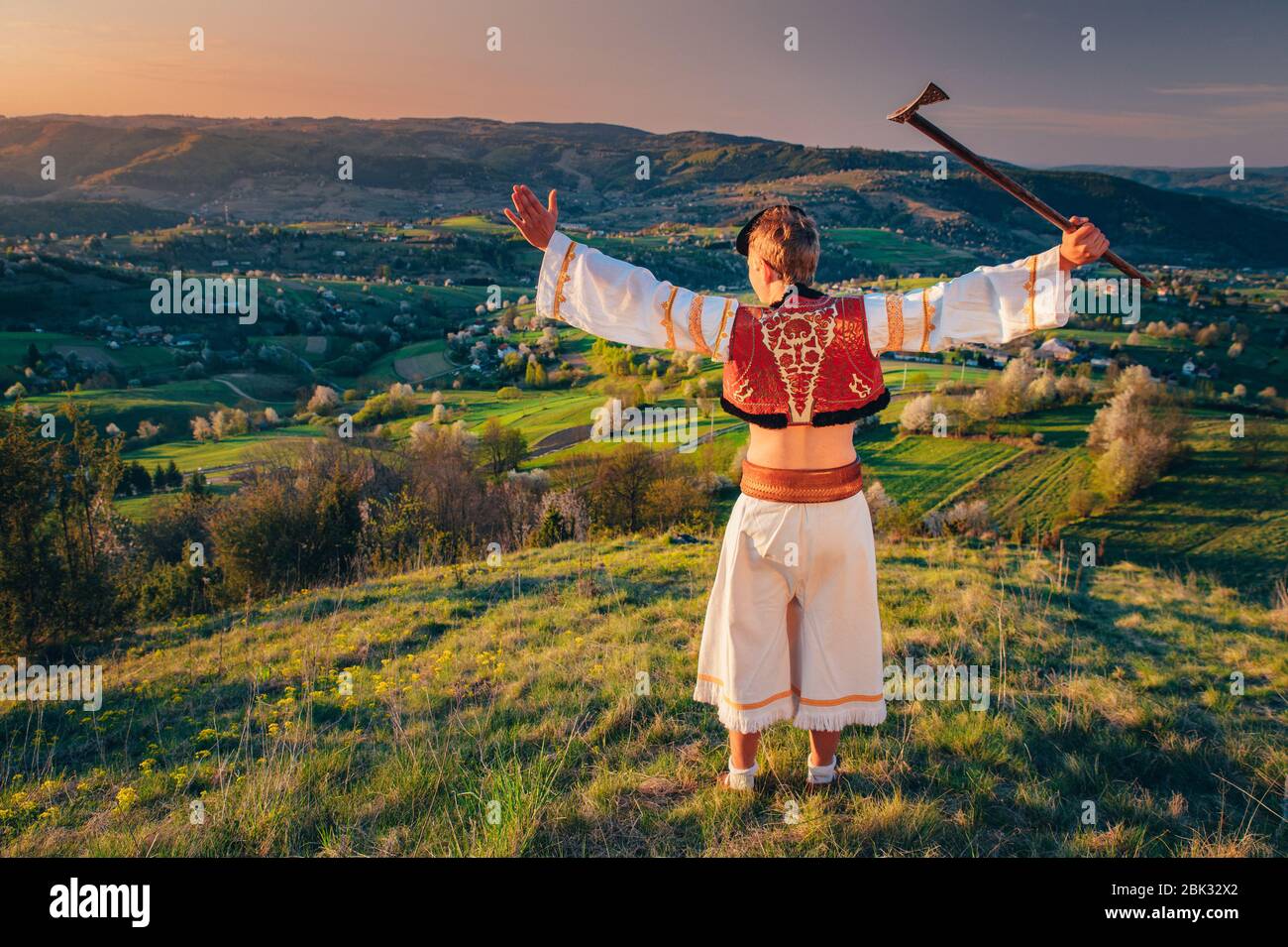 Glücklicher junger Mann mit offenen Armen in traditioneller slowakischer Tracht, der in einer wunderschönen Frühlingslandschaft im Sonnenaufgang steht Stockfoto