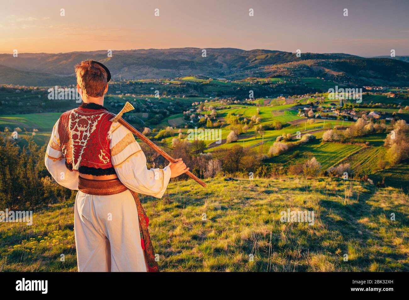 Ein junger Mann in slowakischer Tracht blickt auf die Frühlingslandschaft im Dorf Hrinova in der Slowakei. Aufgehende Sonne und Frühling blühende Bäume in der Stockfoto