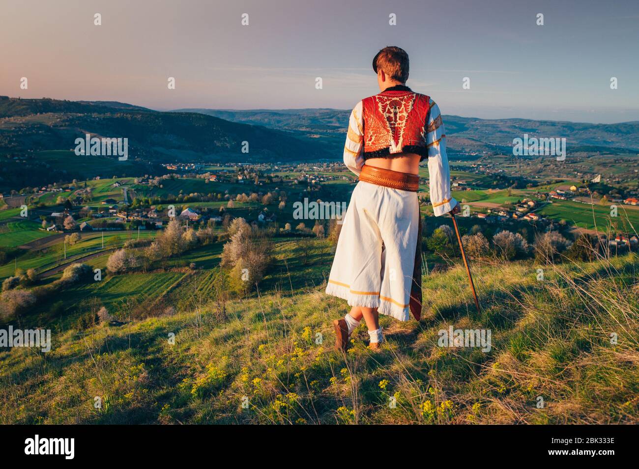 Mann in traditioneller Volkskleid, der das Licht des Sonnenaufgangs in einer wunderschönen, warmen Frühlingslandschaft betrachtet. Ländliche Wiese und Frühlingsblüten Kirschbäume im Hintergrund Stockfoto