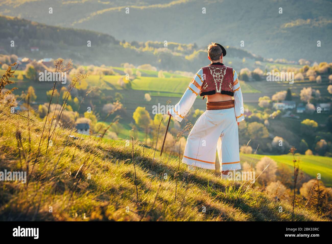 Ein junger Mann in slowakischer Tracht blickt auf die Frühlingslandschaft im Dorf Hrinova in der Slowakei. Aufgehende Sonne und Frühling blühende Bäume in der Stockfoto