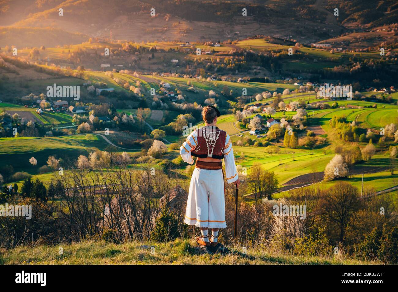 Ein junger Mann in slowakischer Tracht blickt auf die Frühlingslandschaft im Dorf Hrinova in der Slowakei. Aufgehende Sonne und Frühling blühende Bäume in der Stockfoto