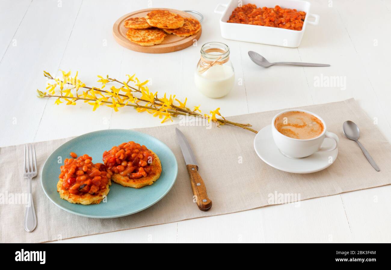 Tisch mit hausgemachten würzigen gebackenen Bohnen mit Speck und Kartoffelbröseln, Tasse Kaffee, Glas Sahne, mit wenigen Zweigen von goldenem Regen verziert. Englisch Stockfoto