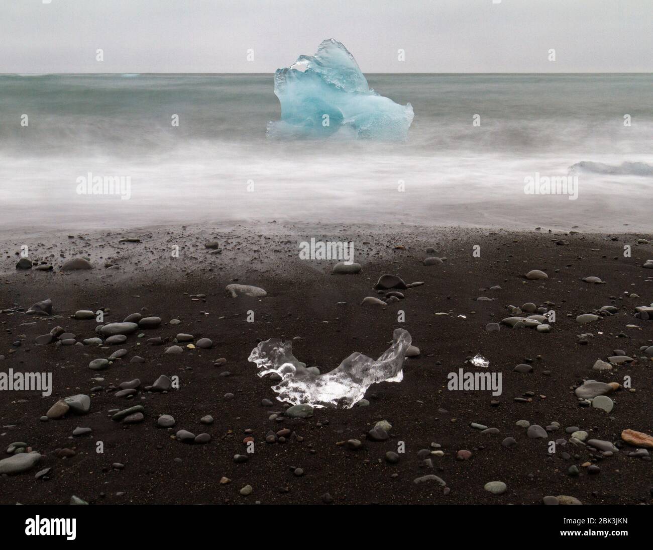 Lange Exposition eines der vielen Eisstücke am Jökulsárlón Diamantenstrand mit einem großen Eisberg im Meer, Südisland. Stockfoto