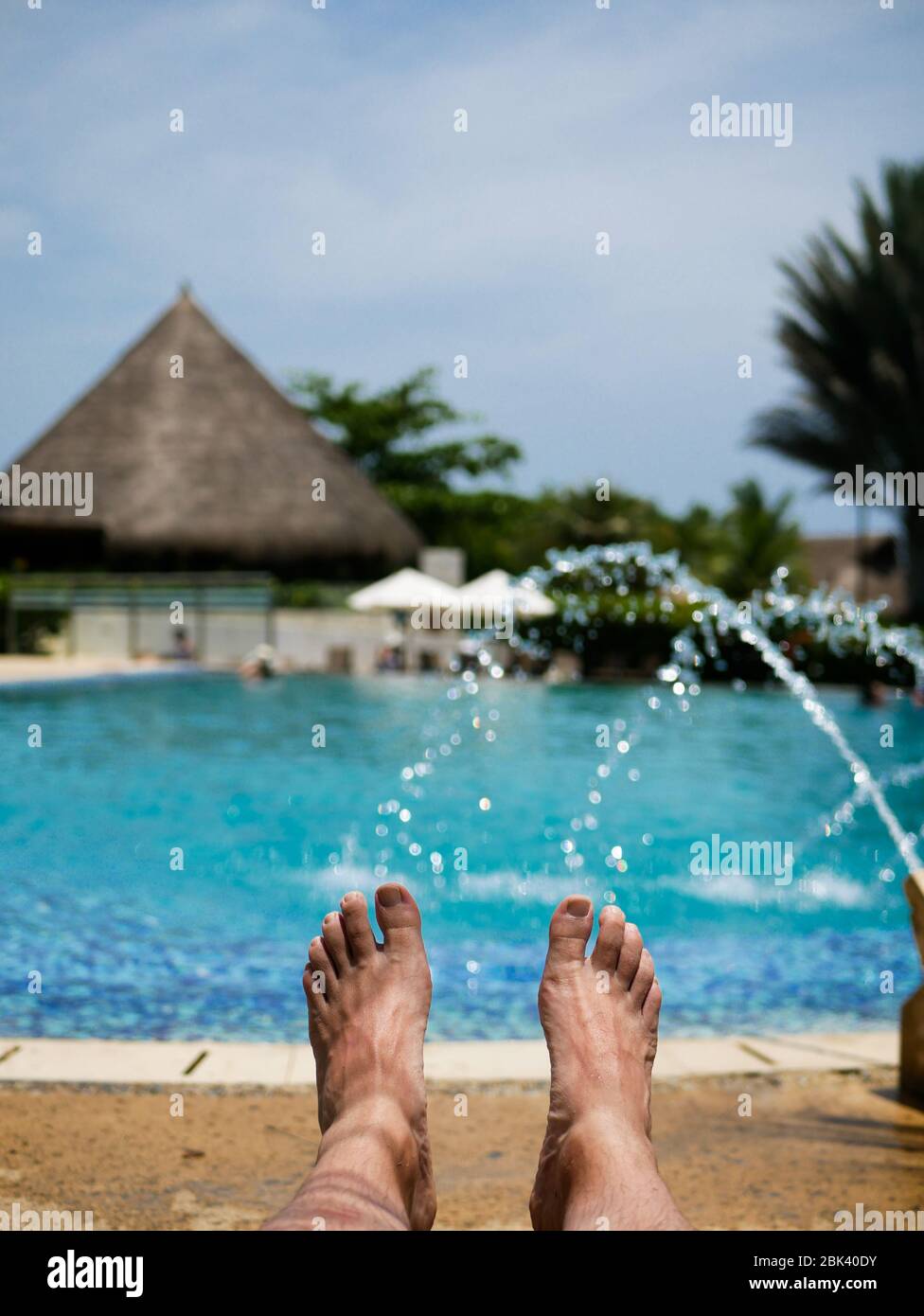 Barfuß Füße eines Mannes, der ein Sonnenbad in einem Pool mit Brunnen und eine Hütte im Hintergrund vertikal Stockfoto