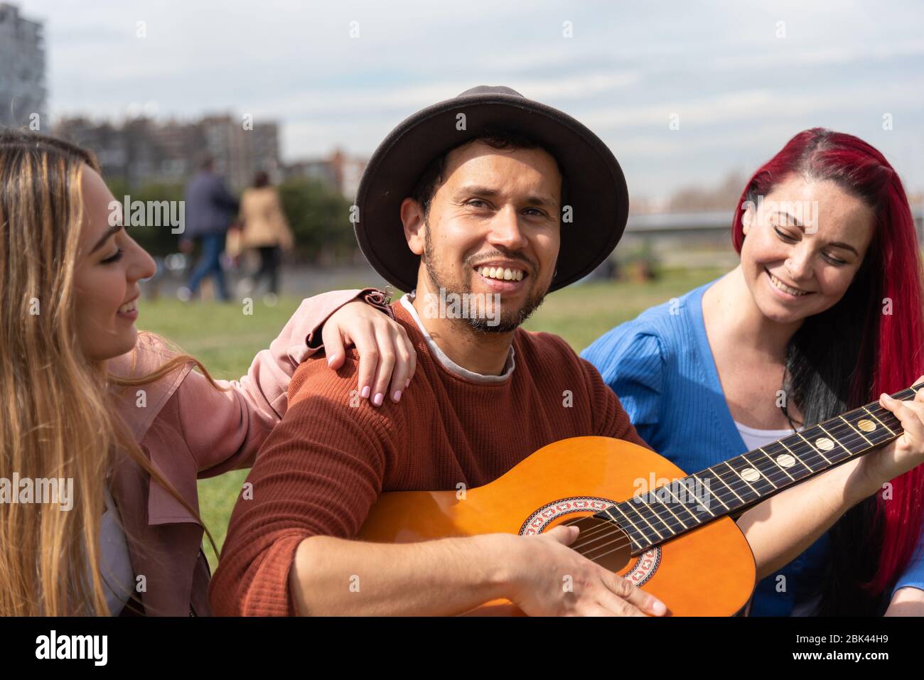 Ein hispanischer Junge in einem Hut spielt neben zwei kaukasischen Mädchen in einem Stadtpark die Gitarre Stockfoto