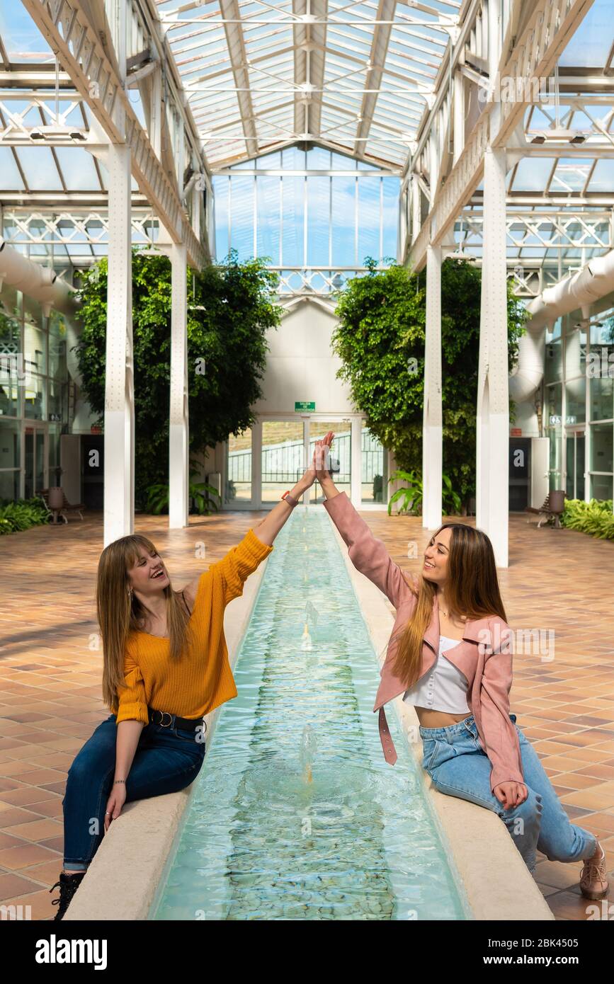 Zwei junge kaukasische Frauen sitzen gerne am Rande eines Brunnens mit Wasser in einem hellen Gewächshaus Stockfoto