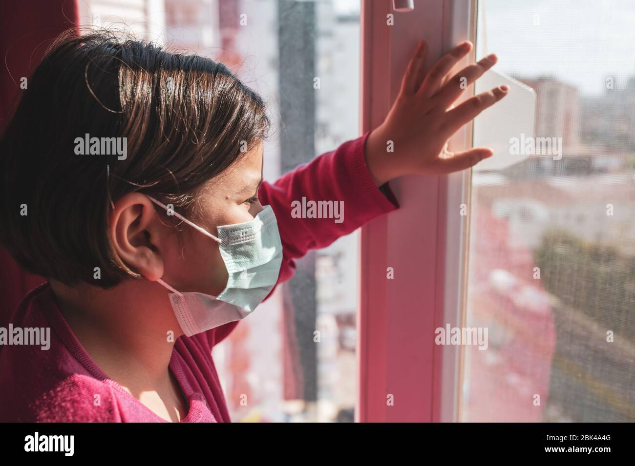 Kleines Mädchen, Kind in medizinischer Maske auf Fenstern, Covid-19 Coronavirus Quarantäne, Hausschule. Bleiben Sie zu Hause. Konzept der Covid-19 Coronavirus Quarantäne Stockfoto