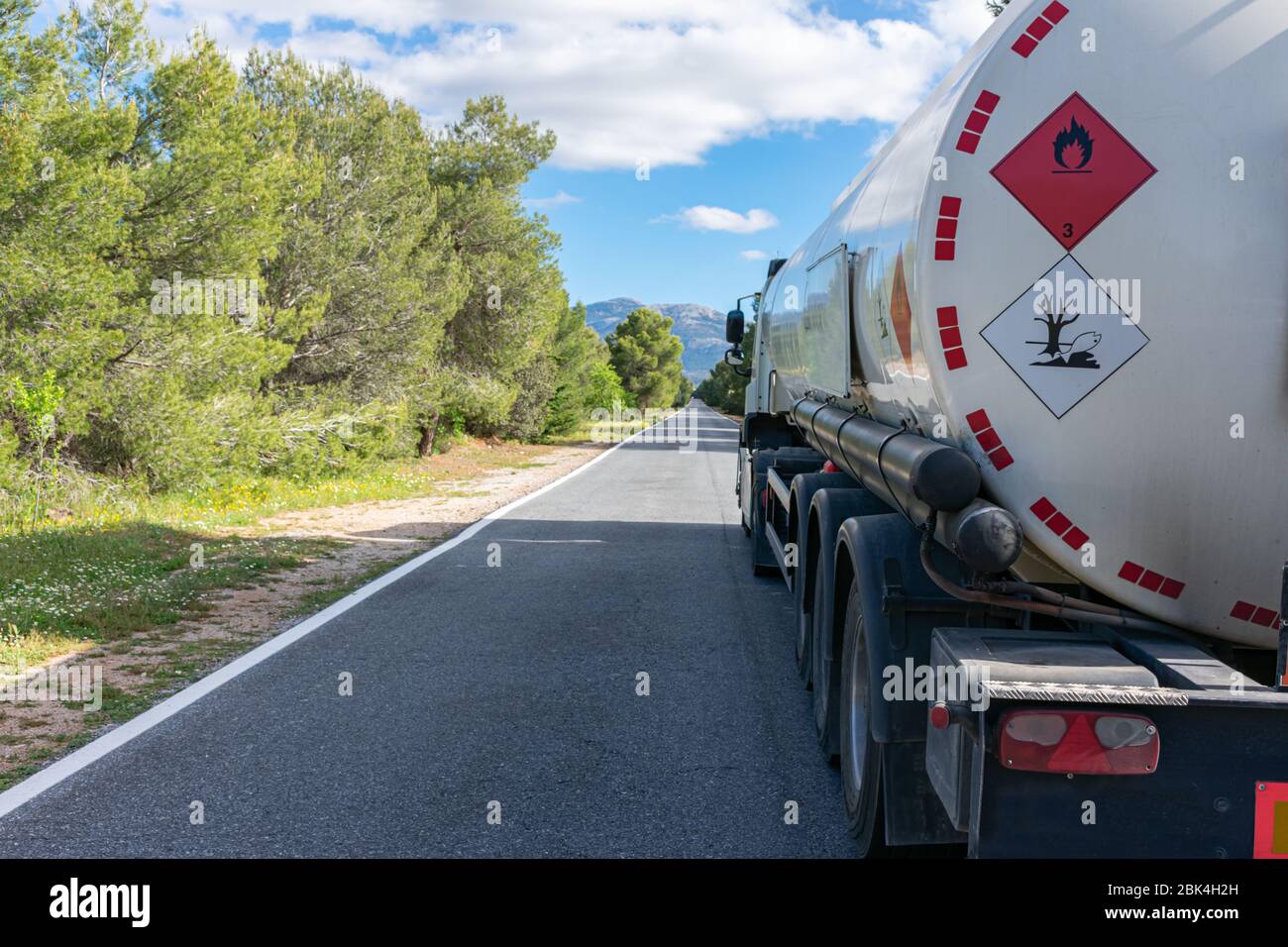 Gefahrenetiketten für brennbare und umweltschädliche Flüssigkeiten auf einem Tankwagen, der auf einer schmalen, geraden Straße mit Bäumen fährt. Stockfoto