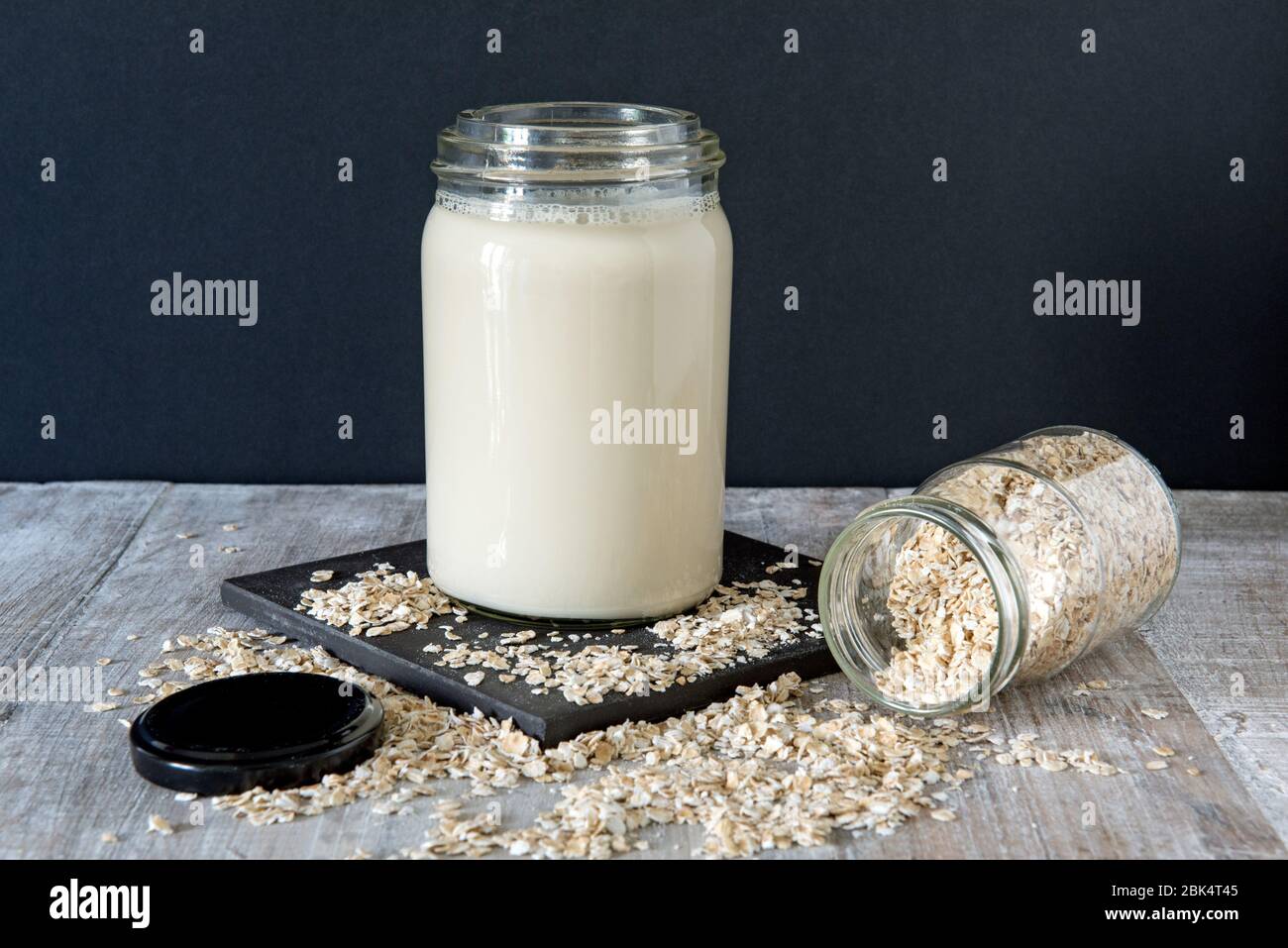 Vegane Hafermilch oder trinken Sie im Glas auf schwarzer Fliese mit verschütteten Hafer und Deckel vor schwarzem Hintergrund links Stockfoto