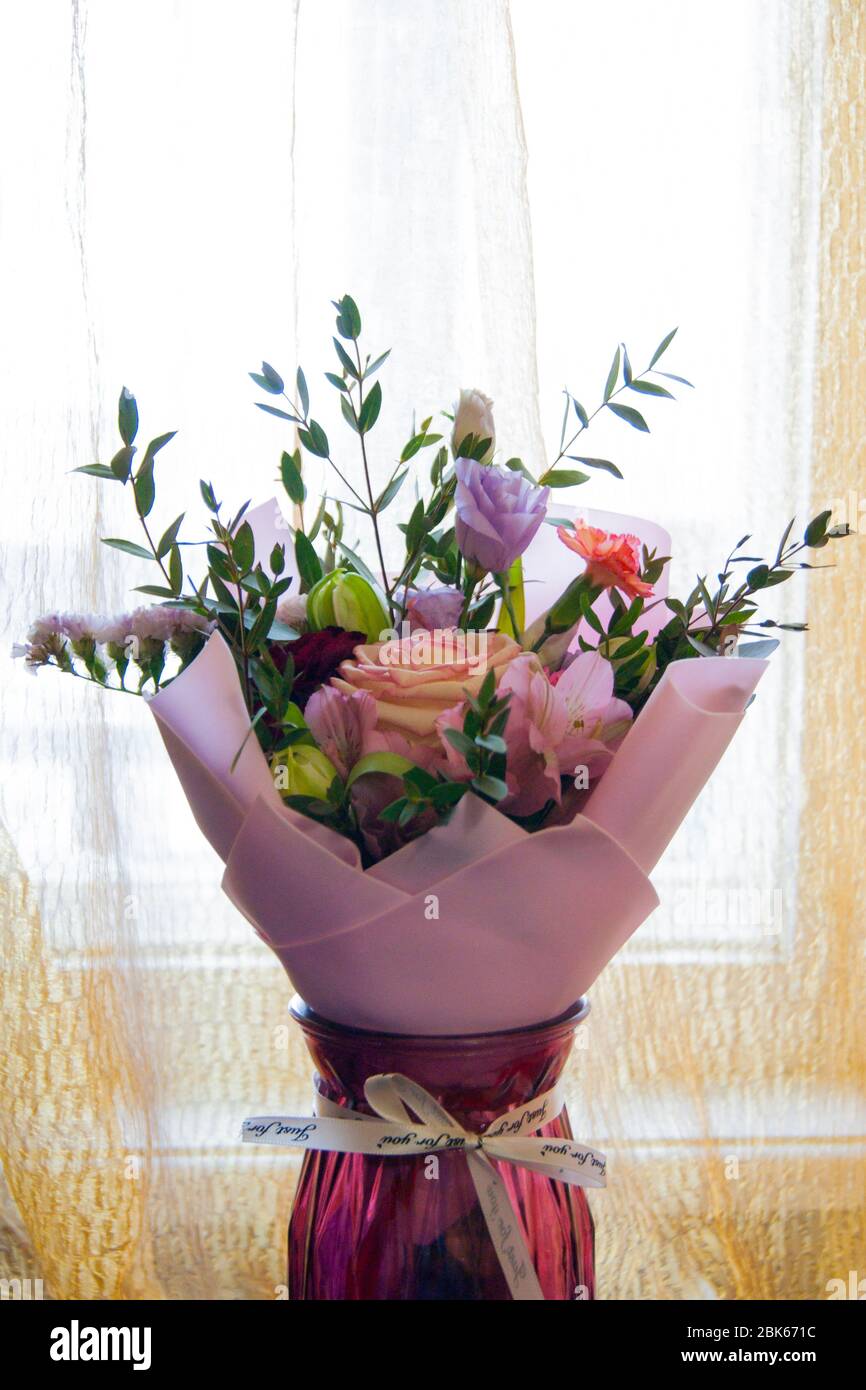 Ein zartes Bouquet aus verschiedenen Blumen in einer hellrosa Umhüllung steht auf einem hellen Hintergrund in einer häuslichen Umgebung. Vertikales Foto. Stockfoto