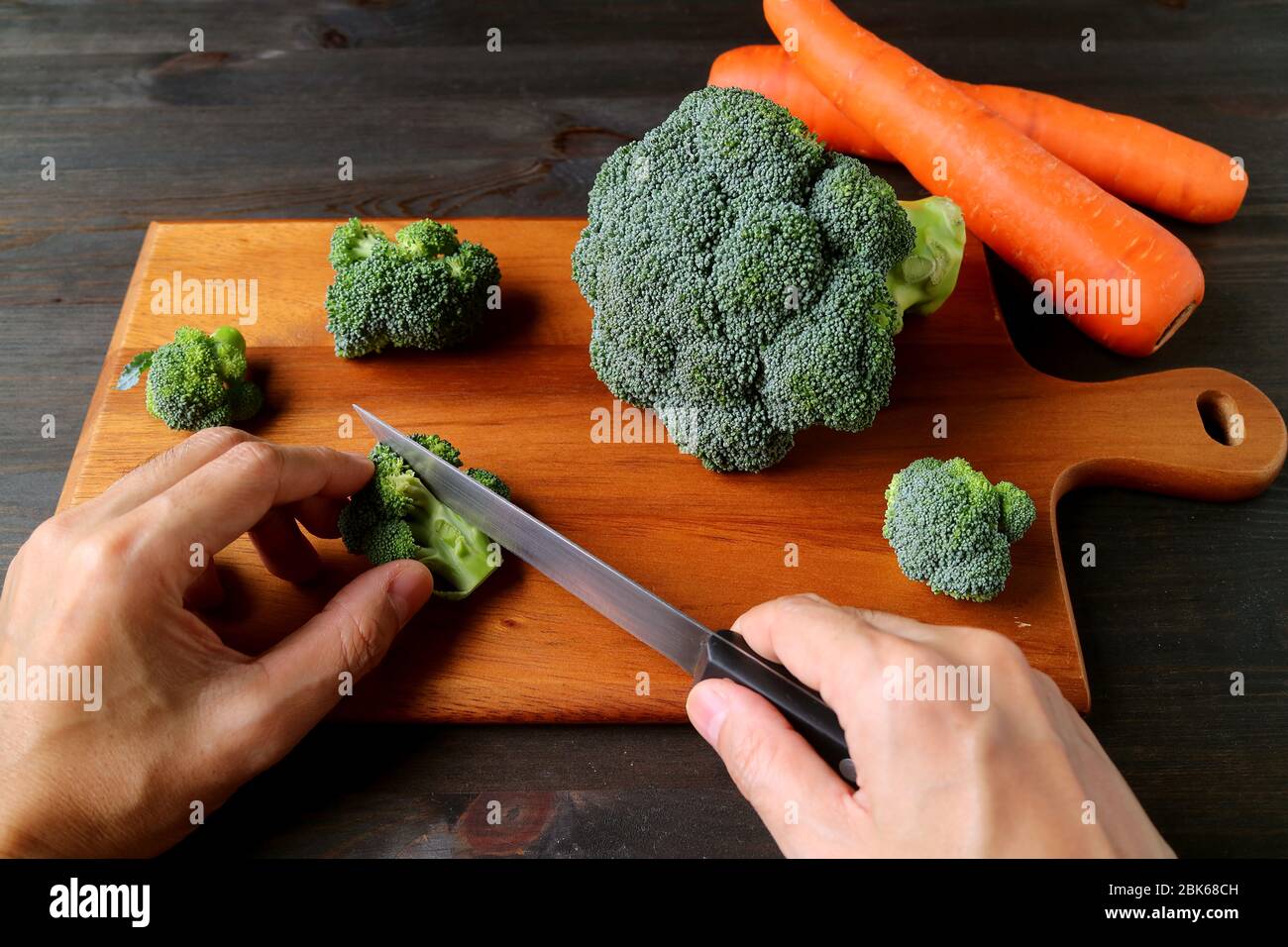 Hand schneiden Roh Broccoli auf Holz Schneidebrett mit verschwommenen Karotten im Hintergrund Stockfoto