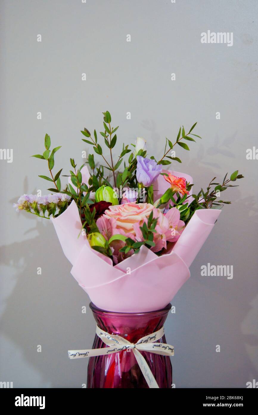 Ein zartes Blumenstrauß verschiedener Blumen in einem hellrosa Wrapper steht in einer weinroten Vase auf grauem Grund. Vertikales Foto. Stockfoto