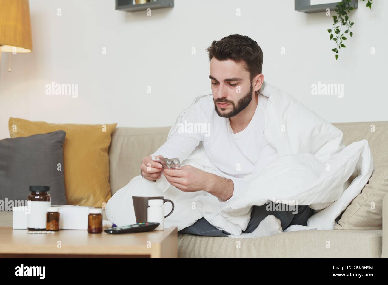 Kranker bärtiger Mann mit hoher Körpertemperatur, der Pillen oder Tabletten während des Aufenthalts zu Hause wegen Coronavirus oder einer anderen Krankheit nimmt Stockfoto